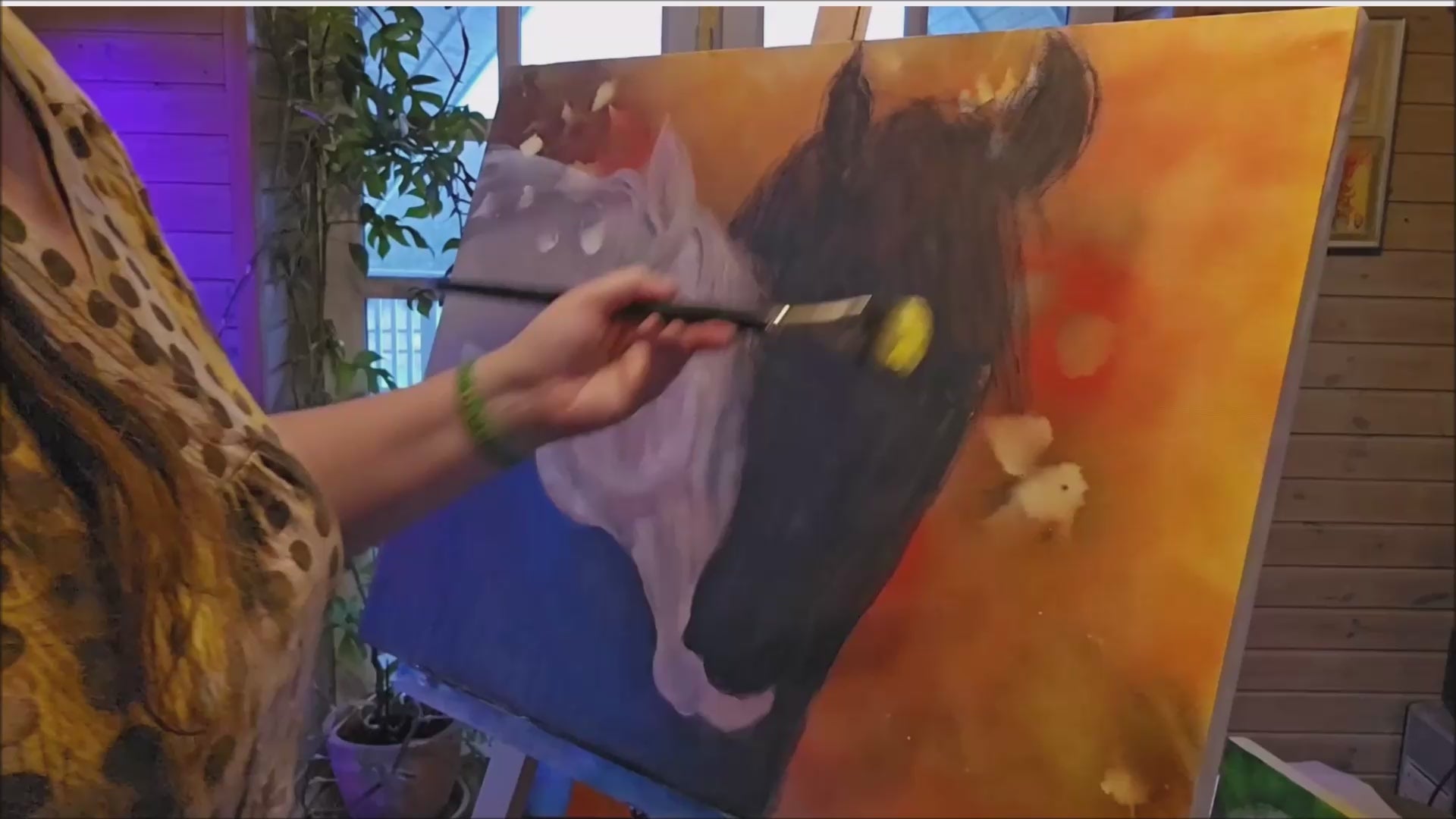 Indlæs video: Kunstneren Anna Maria maler på maleriet Trøst med et tyndt lag olie. Maleriet består af to heste, en sort og en hvid som stikker hovederne sammen på en baggrund i gul, orange og røde nuancer.